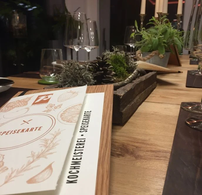 Gedeckter Chef’s Table in der Kochmeisterei mit Weingläsern, Kerzen, Kräutern und Naturdekoration.
