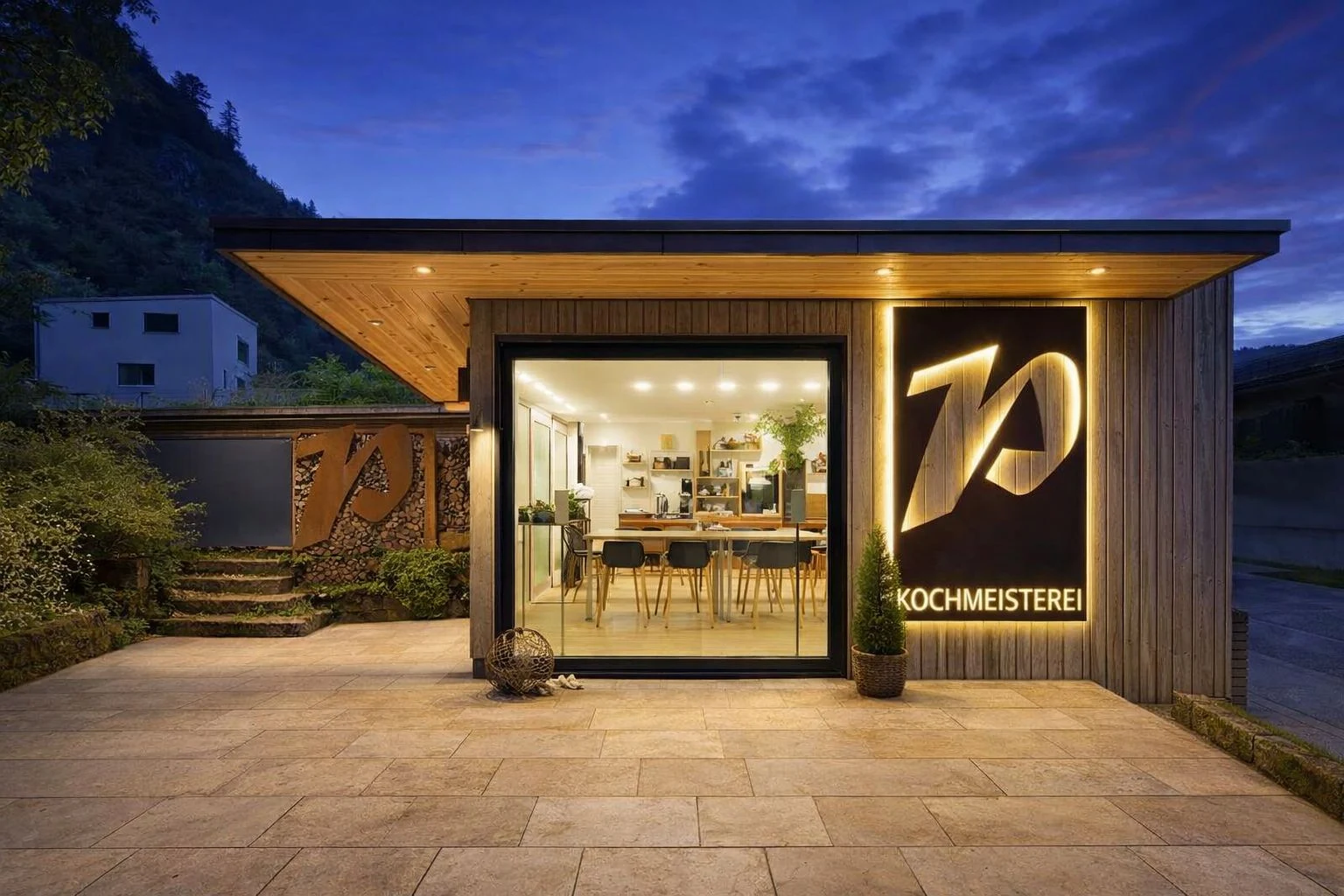 Die Cooking Box der Kochmeisterei in der Abenddämmerung, mit überdachtem Eingang, Terrasse und beleuchtetem Pansi’s-Zeichen.